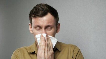 Sick man with red nose sneezes hard wiping with white napkin standing on grey background. Young male suffers from allergy to dust close view