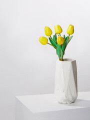 Subject shot of a graceful bouquet of tender yellow tulips in a white ceramic vase. The vase with a bunch of flowers is located on the white table on the white background. 