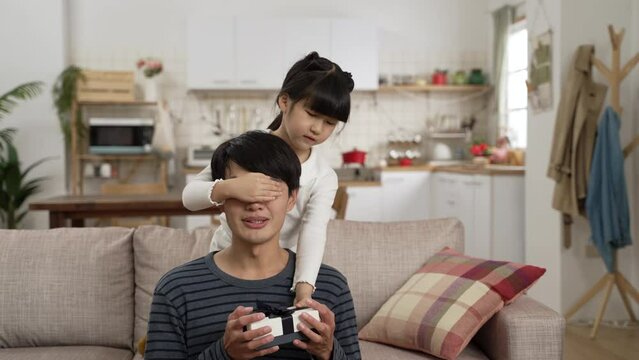 asian girl giving her dad a present and releasing hands covering his eyes from behind on Father’s Day at home. happy man looking at gift with smile while his daughter hugs him