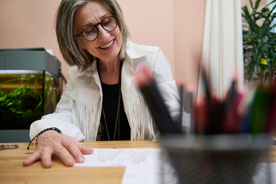 View through blurred colorful pencils holder to a fashion designer, dressmaker, mature elegant Caucasian business woman working at wooden desk, drawing sketches for clothes in her creative workshop