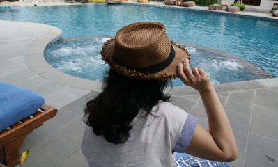 A long-haired female tourist in a white short-sleeved shirt raises her right hand and grabs the tip of a dark brown straw hat as she sits by a pool of bright blue water during her vacation.
