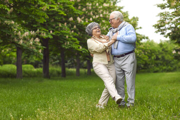 Naklejka na ściany i meble Senior man and woman are dancing on the green grass in the park among the trees. A man holds a woman, wear glasses, and a scarf, happy family life. Walk in the fresh air.