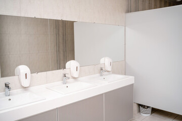 The interior of a public toilet. Row of wash basins with metal faucets on a marble slab, liquid soap dispensers, a long mirror on a gray wall