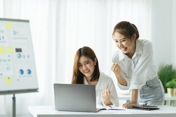 Two pretty young Asian businesswoman sitting at desk with laptop doing paperwork together discussing project financial report. Corporate business collaboration concept.