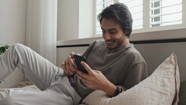 Medium long of young cheerful Middle Eastern man in casual clothes lying on bed, using smartphone in morning, scrolling feed, smiling