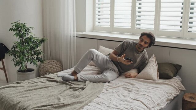 Young cheerful Middle Eastern man in loungewear lying on bed, using smartphone in morning in minimalist house, scrolling feed