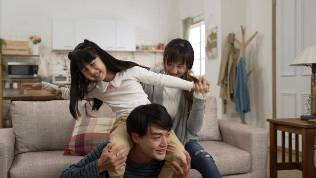 slow motion of happy asian little girl having fun playing on dad’s shoulders in the living room at home. the mom is laughing and holding her daughter’s arms