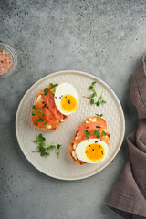 Sandwich with delicious toppings smoked salmon, eggs, herbs and microgreens radish, black sesame seeds over white plate on gray concrete table background. Healthy open sandwich superfood. Top view.