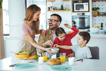 Happy family of four at the table, mother serves them breakfast. Everyone is happy and smiling, parents enjoy spending time with their children. 
