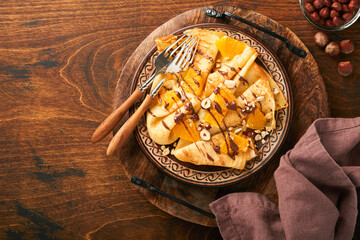 Crepes suzette with oranges and chocolate. Thin crepes with chocolate spread, hazelnuts and orange slices fruit in plate for breakfast on old dark wooden rustic table background. Copy space. Top view