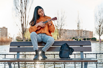 young latina playing ukulele in a city park. woman sitting on a bench practicing with her musical instrument. - obrazy, fototapety, plakaty
