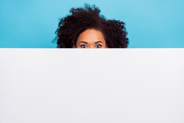 Photo of young person eyes peeking behind empty space proposition poster isolated on blue color...