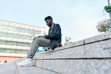 Obraz premium side view of an african american man working with his laptop sitting on a staircase in the city, wearing casual clothes.