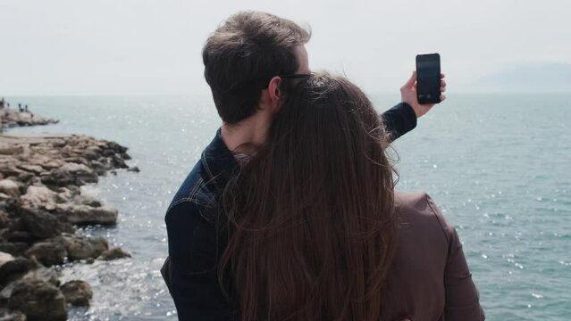 Back view of oving couple of travelers a guy and a girl have a video call conversation on the phone in a picturesque place on the ocean. Young man and woman take a selfie on the beach. High quality 4k