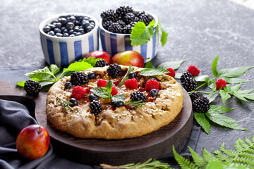 Whole-grain galette with plums and berries on dark background, top view