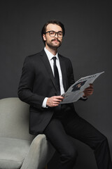 businessman in suit and glasses leaning on armchair and holding business newspaper on dark grey.