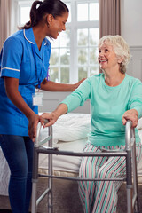 Senior Woman At Home Using Walking Frame Being Helped Out Of Bed By Female Care Worker In Uniform