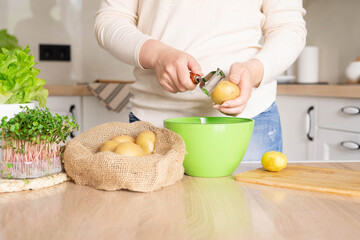 Woman peels potatoes with a vegetable peeler standing in the kitchen.