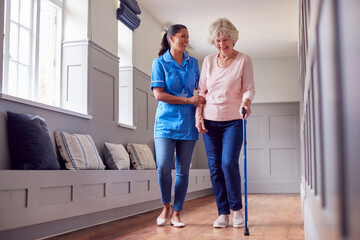 Senior Woman At Home Using Walking Stick Being Helped By Female Care Worker In Uniform