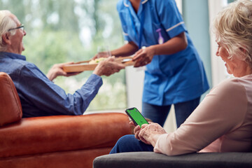 Close Up Of Senior Woman Using Mobile Phone As Carer Brings Meal To Senior Man In Lounge At Home 