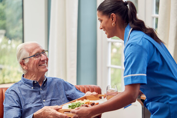 Female Care Worker In Uniform Bringing Meal On Tray To Senior Man Sitting In Lounge At Home 