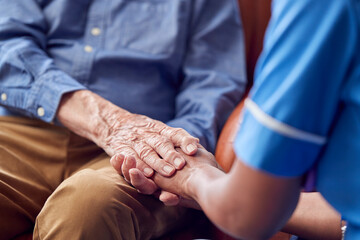 Close Up Of Female Care Worker In Uniform Holding Hands Of Senior Man Sitting In Care Home Lounge 