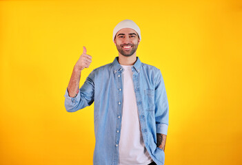 Showing Like. Studio shot of smiliing handsome man in jeans and white hat on yellow background. Tatoo and beard. Happy concept.