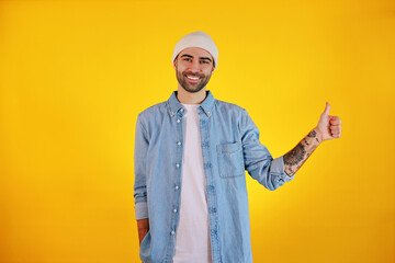 Showing Like. Studio shot of smiliing handsome man in jeans and white hat on yellow background. Tatoo and beard. Happy concept.