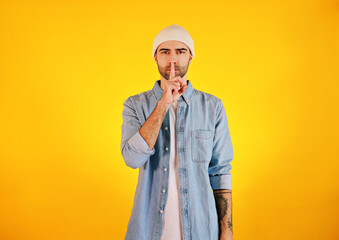 Showing hush sing. Studio shot of smiliing handsome man in jeans and white hat on yellow background. Tatoo and beard.