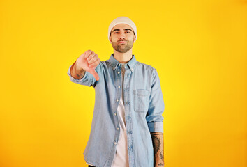 Showing dislike. Studio shot of smiliing handsome man in jeans and white hat on yellow background. Tatoo and beard.