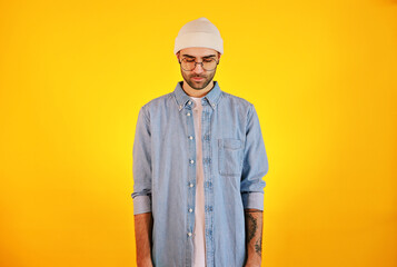 Studio shot of  handsome man in jeans , glasses and white hat on yellow background. Thinking concept. Tatoo and beard.