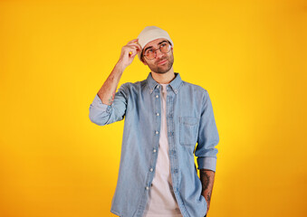 Studio shot of  handsome man in jeans , glasses and white hat on yellow background. Thinking concept. Tatoo and beard.