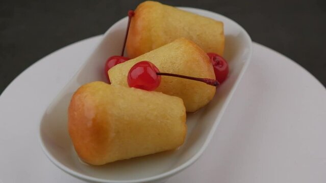 Neapolitan Rum baba on a white plate with a cocktail cherry. Small yeast cakes soaked in rum syrup on a black background. Traditional Italian pastry. Rotation, slow motion. Close-up, selective focus.