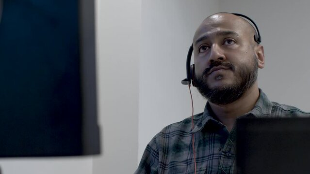 Bald Bearded UK Asian Male Wearing Headset Swivelling On Chair Waiting For Call. View Behind Monitor. Low Angle, Locked Off