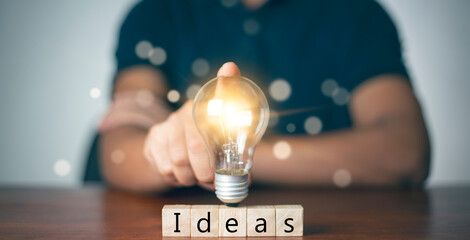 A man's hand touches a light bulb on a wooden block with Word Ideas, innovative and inspiring new...