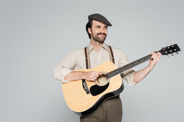 smiling man with mustache in retro style clothing and hat playing acoustic guitar isolated on grey.