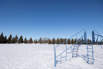 Playground equipment in a snow covered field with an evergreen tree line in the background and a bright blue sky