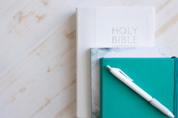 White Christian bible with teal journal and white pen and devotional on a white wood background...