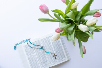 Open bible and blue rosary prayer beads with spring tulips on a white background with copy space