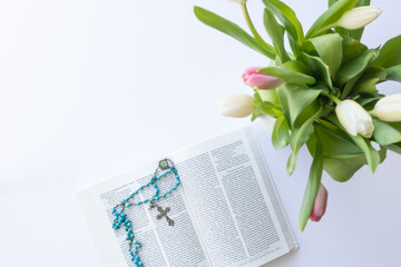 Open Christian bible with blue rosary prayer beads laying on a white desk with a vase of pink and...