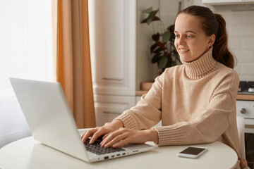 Image of woman with dark hair wearing beige sweater sitting in kitchen and working on laptop and typing on keyboard, expressing positive emotions.