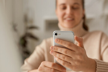 Portrait of smiling woman with blurred face wearing beige sweater using smartphone, reading news or typing messages, expressing positive emotions.