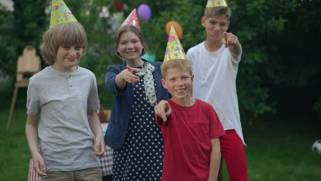Group of children and teenagers teasing pointing at camera laughing standing in summer spring park. Portrait of Caucasian boys and girl mocking posing in slow motion at birthday party outdoors