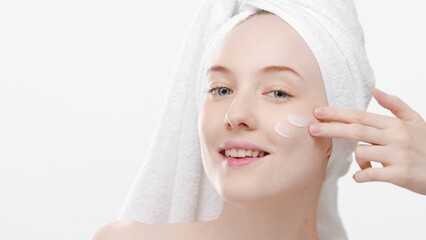 Pretty slim Caucasian young woman with a towel on her head applies face care cream on cheek on white background | Face care cream commercial