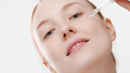 Big close-up shot of young slim ginger European woman drips face serum from dropper on her cheek on white background | Skin care serum commercial