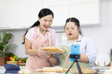 mother and down syndrome teenage girl or her daughter showing bread on basket and live streaming online via smartphone on tripod in a kitchen