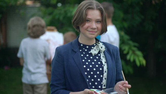 Positive charming Caucasian teenage girl posing with painting palette in slow motion as group of blurred friends painting on easel at background. Portrait of talented inspired teenager smiling