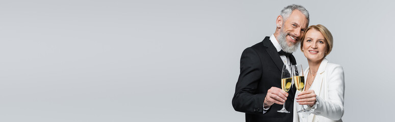 Positive mature couple in wedding clothes holding champagne and looking at camera isolated on grey, banner.