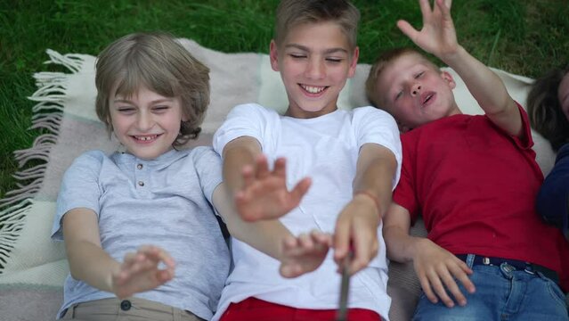 Cheerful friends laughing waving at smartphone video chat lying on weekend picnic outdoors. Happy relaxed Caucasian boys and girl having fun enjoying leisure in spring summer park. Friendship and joy