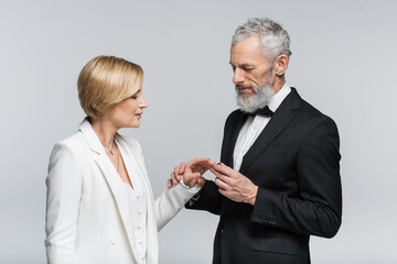 Mature groom wearing wedding ring on finger of bride isolated on grey.